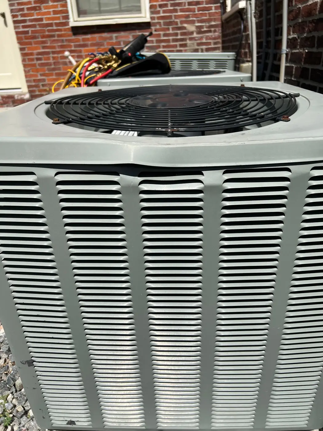Outdoor AC condenser beside a brick house with service hoses laid on top