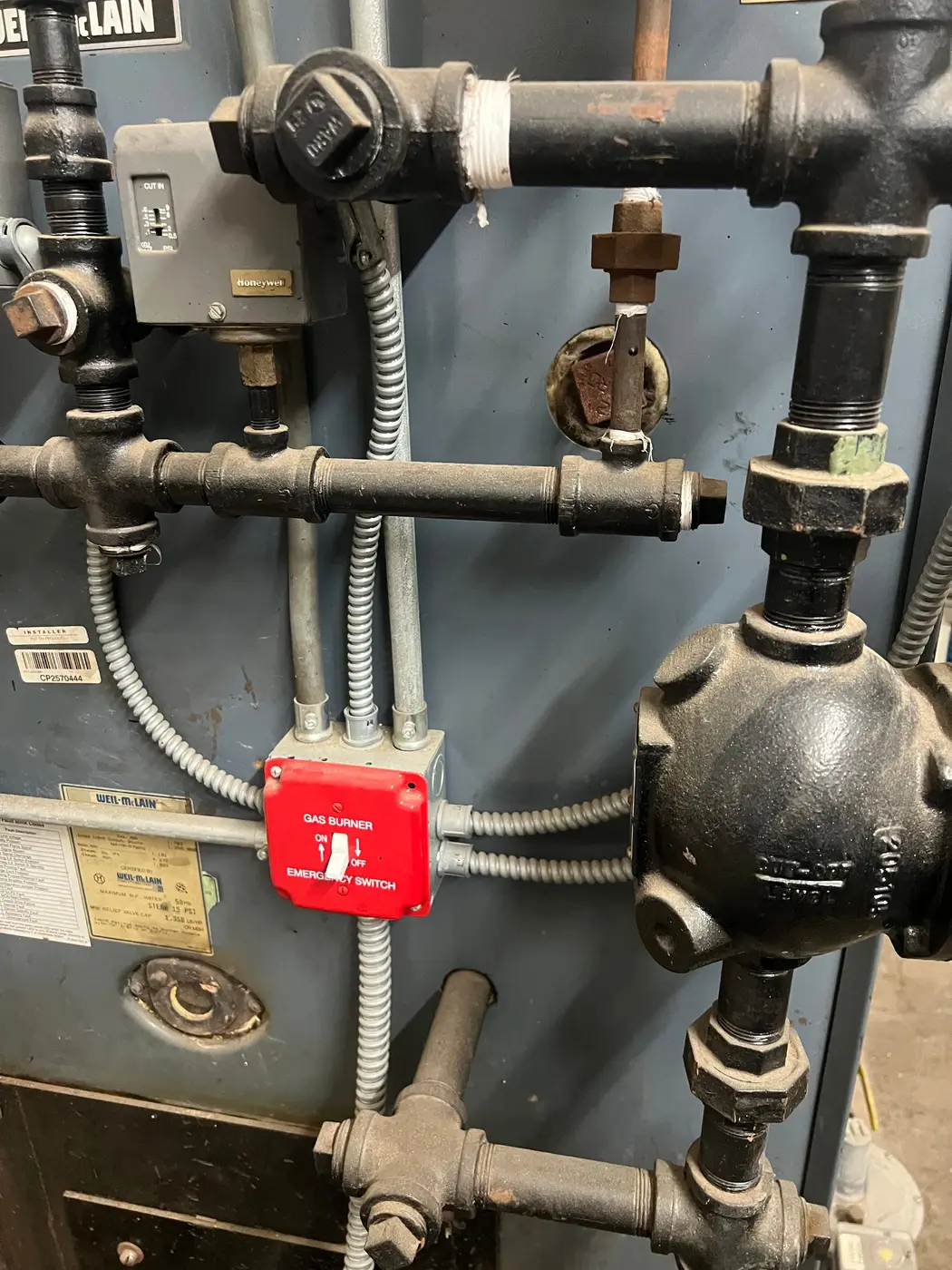 Burner emergency switch, controls and black iron piping on the front of a boiler