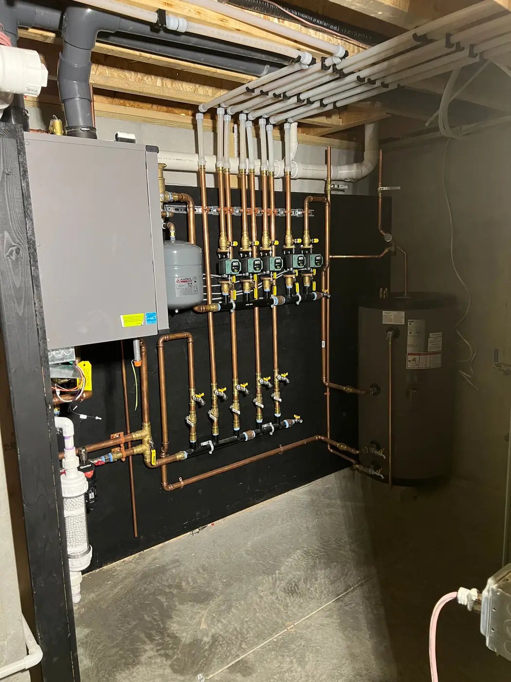 Wide view of a basement boiler installation: wall-hung boiler, zone circulators and indirect tank on a black backboard