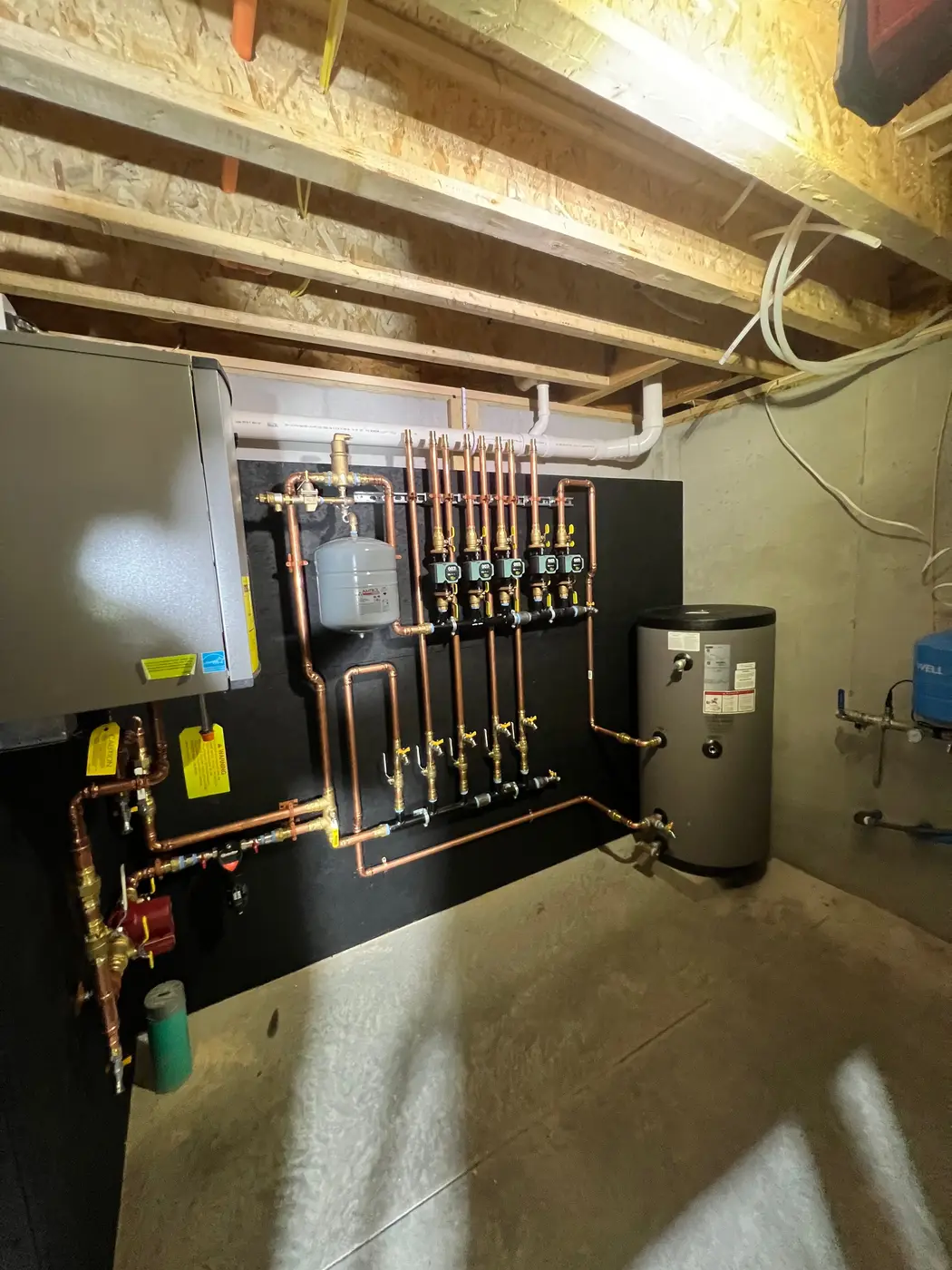 Wall-hung boiler with copper zone piping, circulator pumps and an indirect water heater tank on a black backboard in a basement
