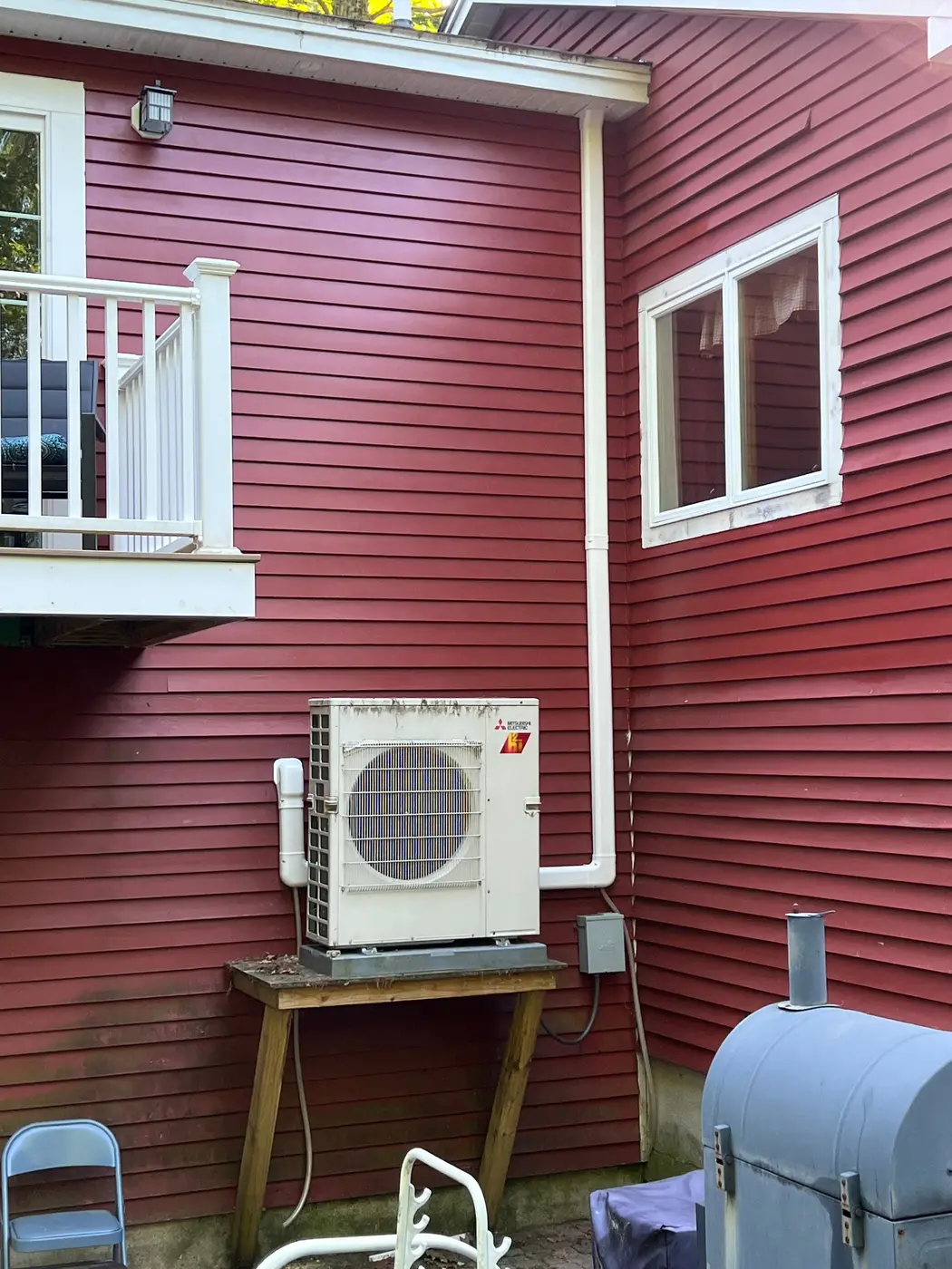 Heat pump outdoor unit on a wooden stand beside a red clapboard house, with a line set cover running up the wall