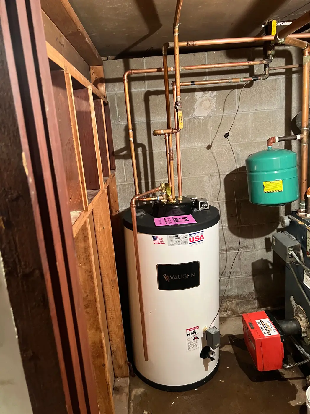 Water heater tank with copper piping and an expansion tank in a basement closet, next to a boiler burner